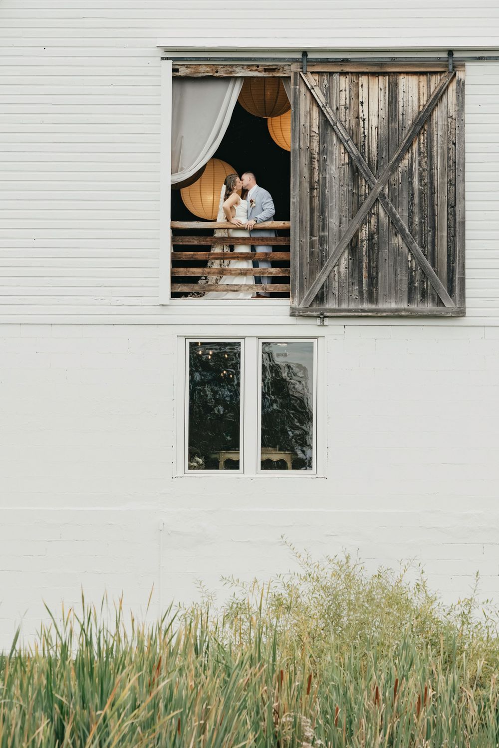 A couple embraces in a rustic barn window with white walls and tall grass growing below.