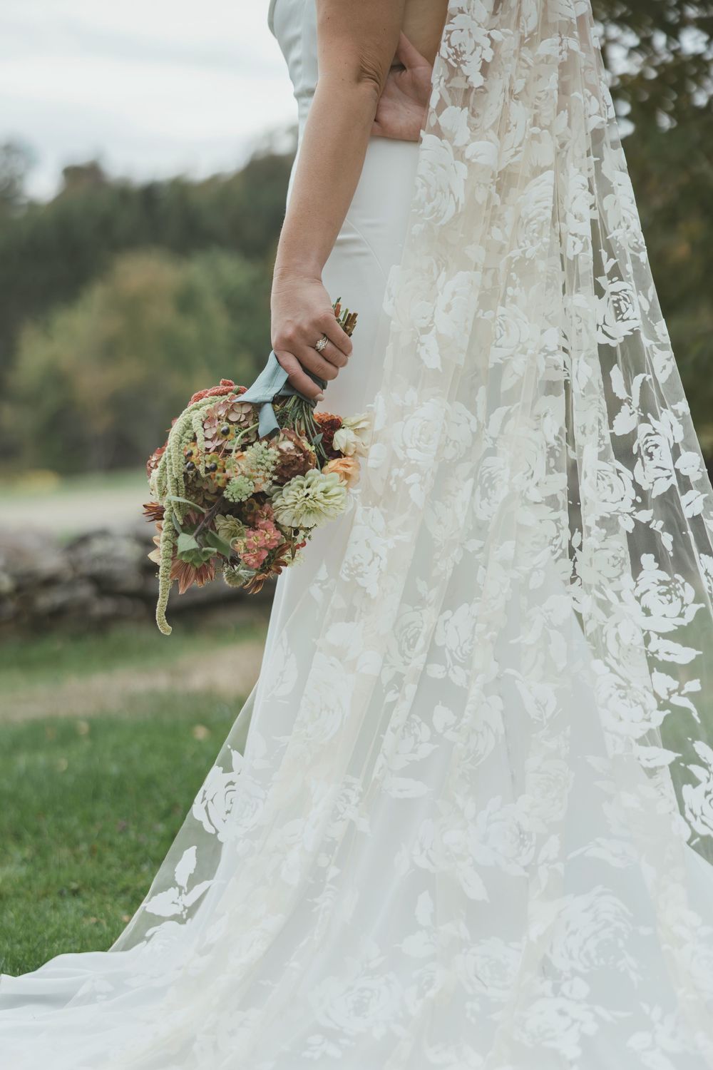 Close-up of a bride holding a rustic bouquet, wearing a detailed lace wedding gown and veil against an outdoor backdrop.