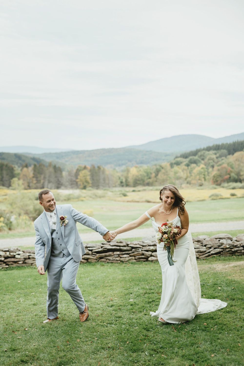 A playful wedding couple runs across a green field with mountains and stone walls in the background.