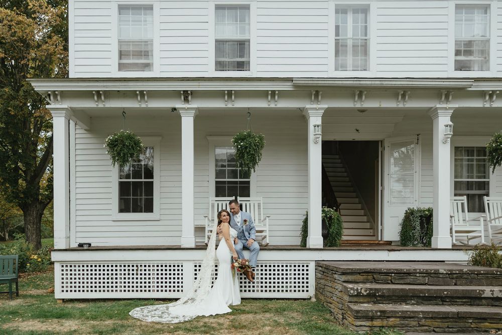 A bride in a white wedding dress poses in front of a classic white farmhouse with a wraparound porch.