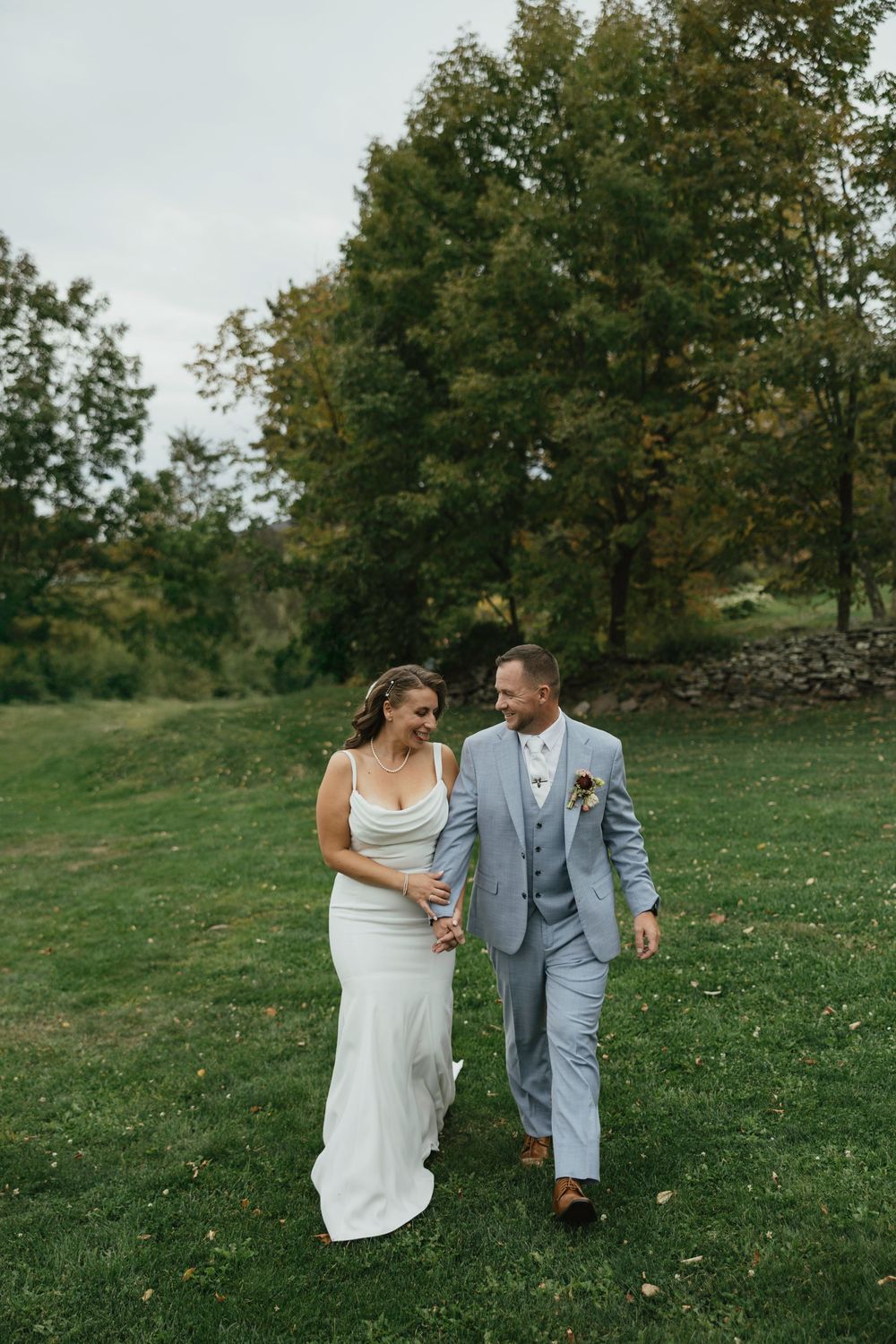 A couple in wedding attire laughing together while walking on a grassy hill with trees in the background.