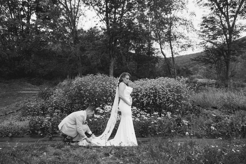 A black and white wedding photograph showing a dress being adjusted in a garden setting with flowering bushes and trees.