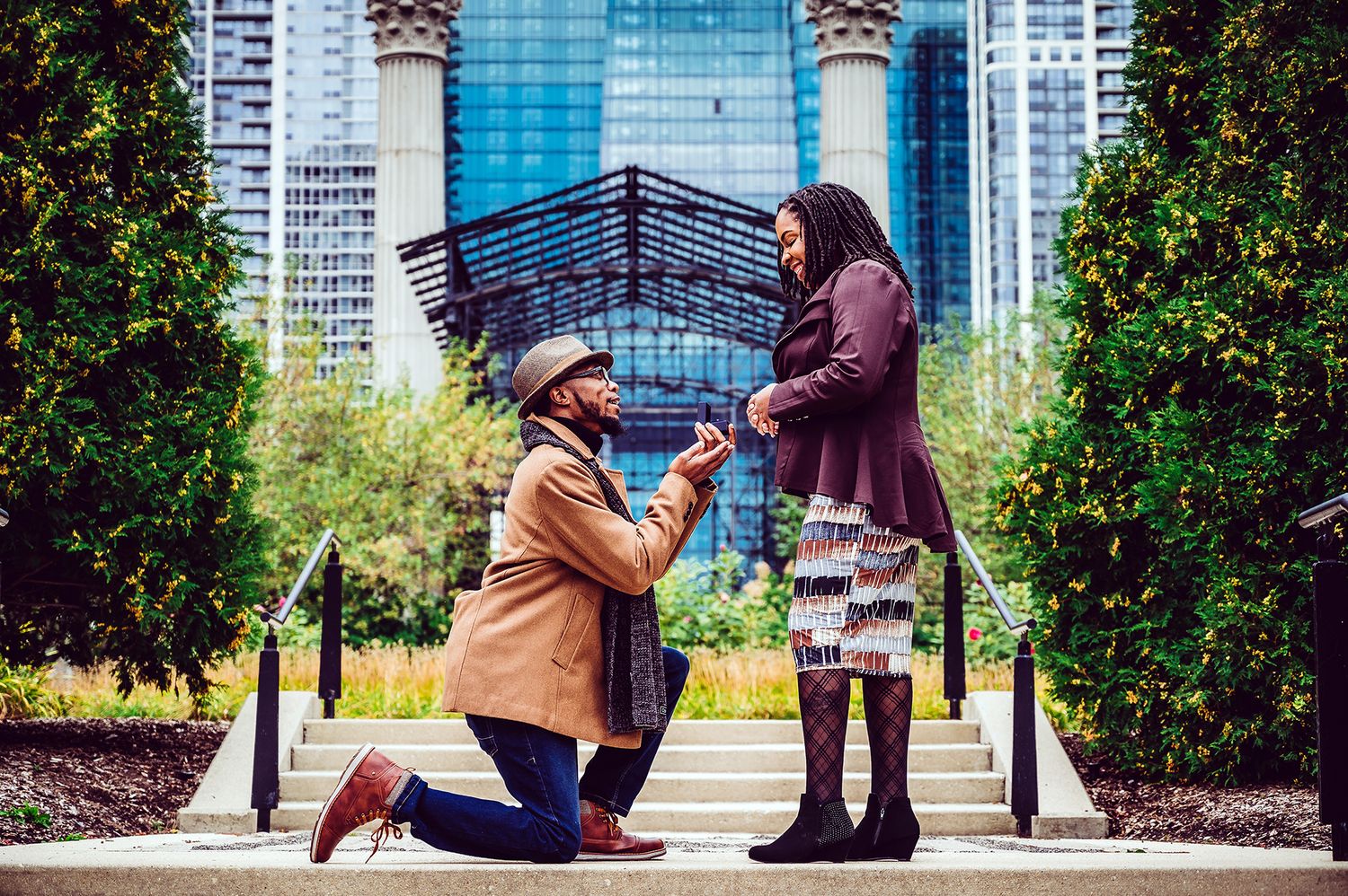 A romantic proposal scene in front of a modern glass building with lush greenery framing the moment.