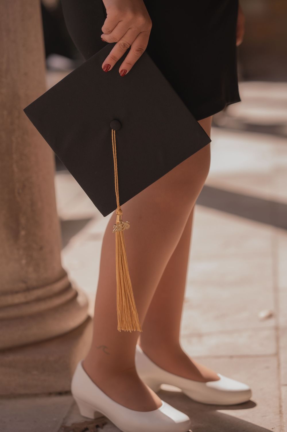 Close-up of a graduate holding a black mortarboard cap with gold tassel, wearing white heels.