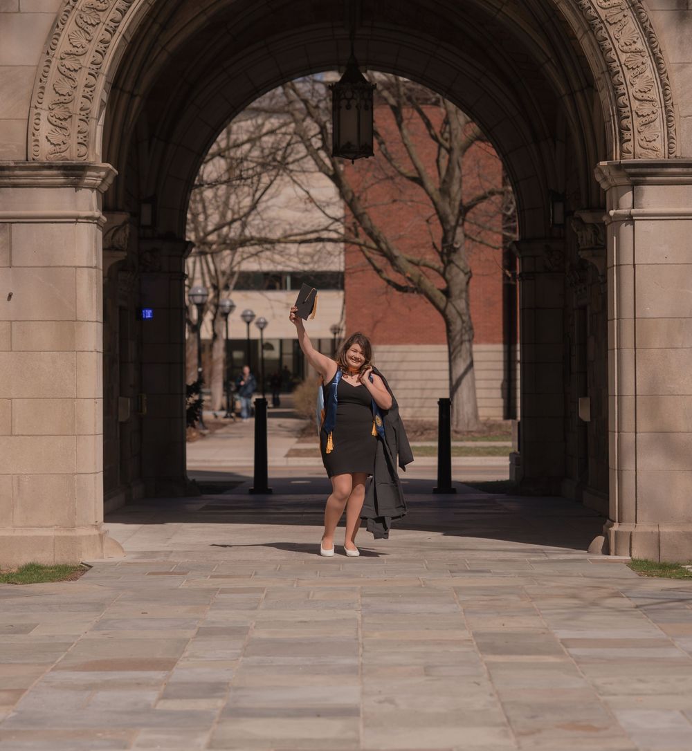 Graduate in cap and gown celebrating under a stone archway on a university campus.