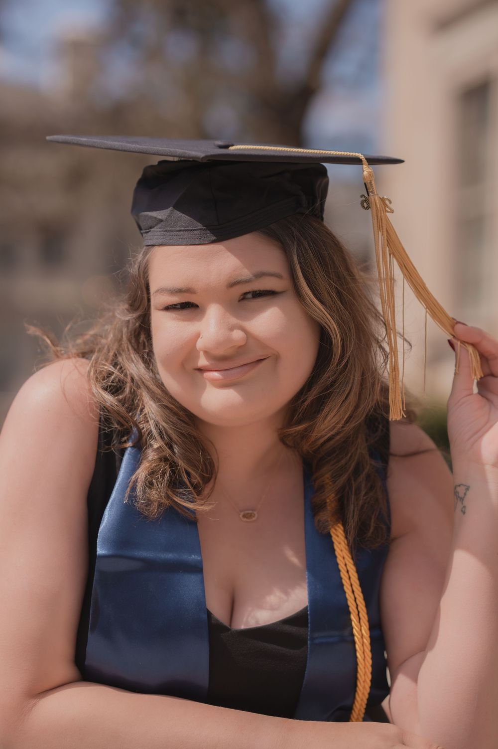 Smiling graduate woman in blue cap and gown holding tassel outdoors on a sunny day.