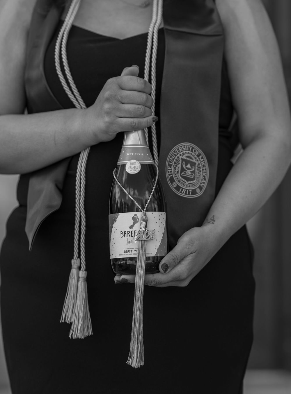 Graduate holding Barefoot sparkling wine bottle and academic sash with honor cords in black and white photo.