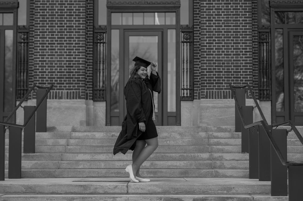 Graduate in black cap and gown walking down steps of a brick building in black and white photo.
