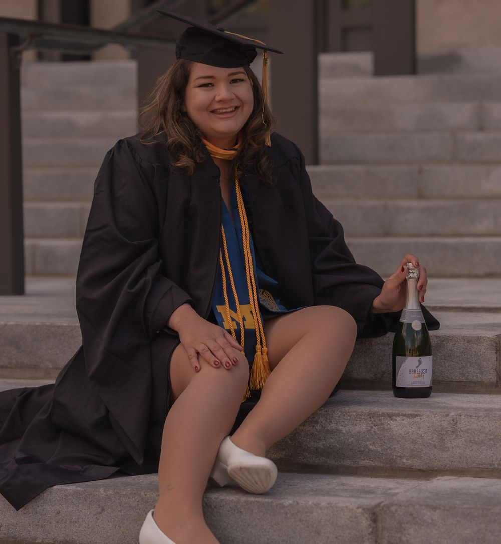 Smiling graduate in black cap and gown sitting on steps holding champagne bottle with gold honor cords.