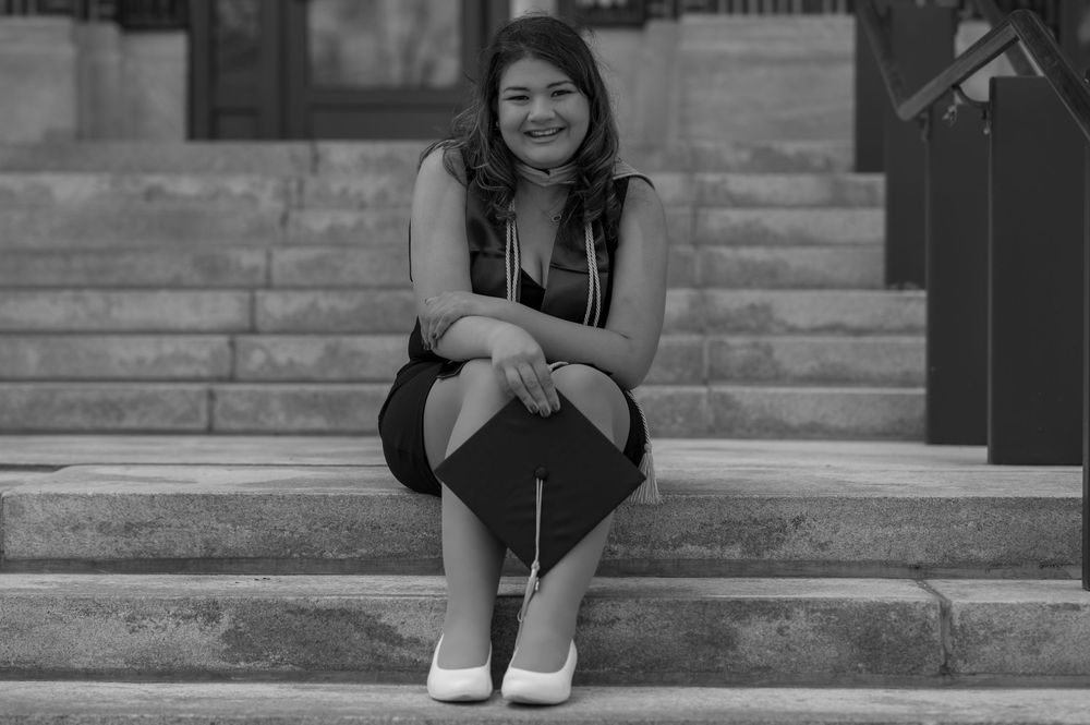Young woman in graduation cap and dress smiling on steps in black and white photo.