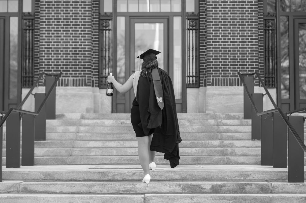 Graduate in cap and gown walks up brick building steps holding diploma in black and white photo.
