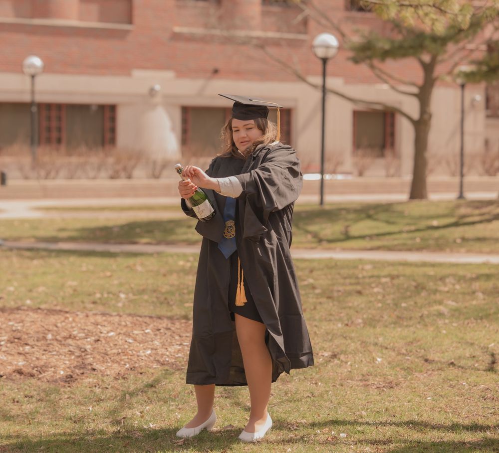 Graduate in cap and gown celebrating outdoors, popping a champagne bottle on a college campus lawn.