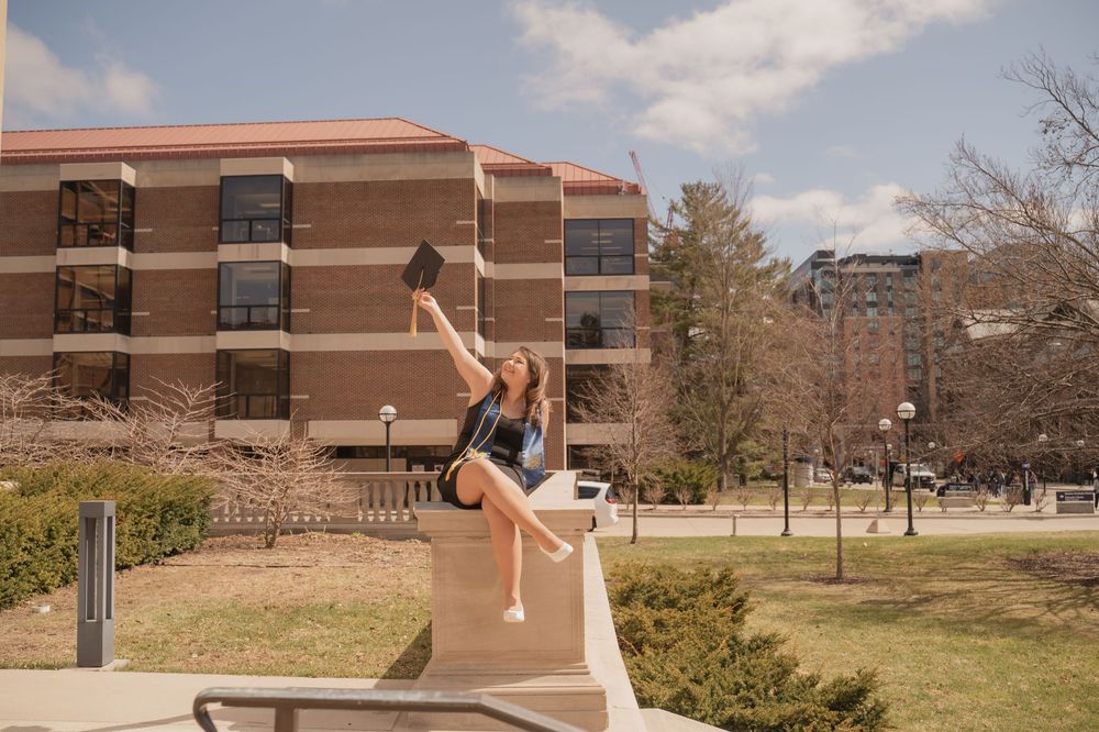 Young woman in cap and gown celebrates graduation outdoors on a sunny college campus with brick buildings.
