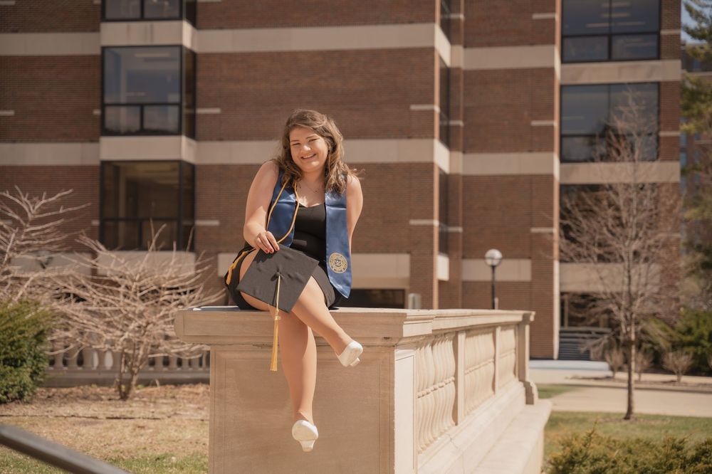 A smiling student sits on a stone wall outside a brick university building, wearing a backpack on a sunny day.