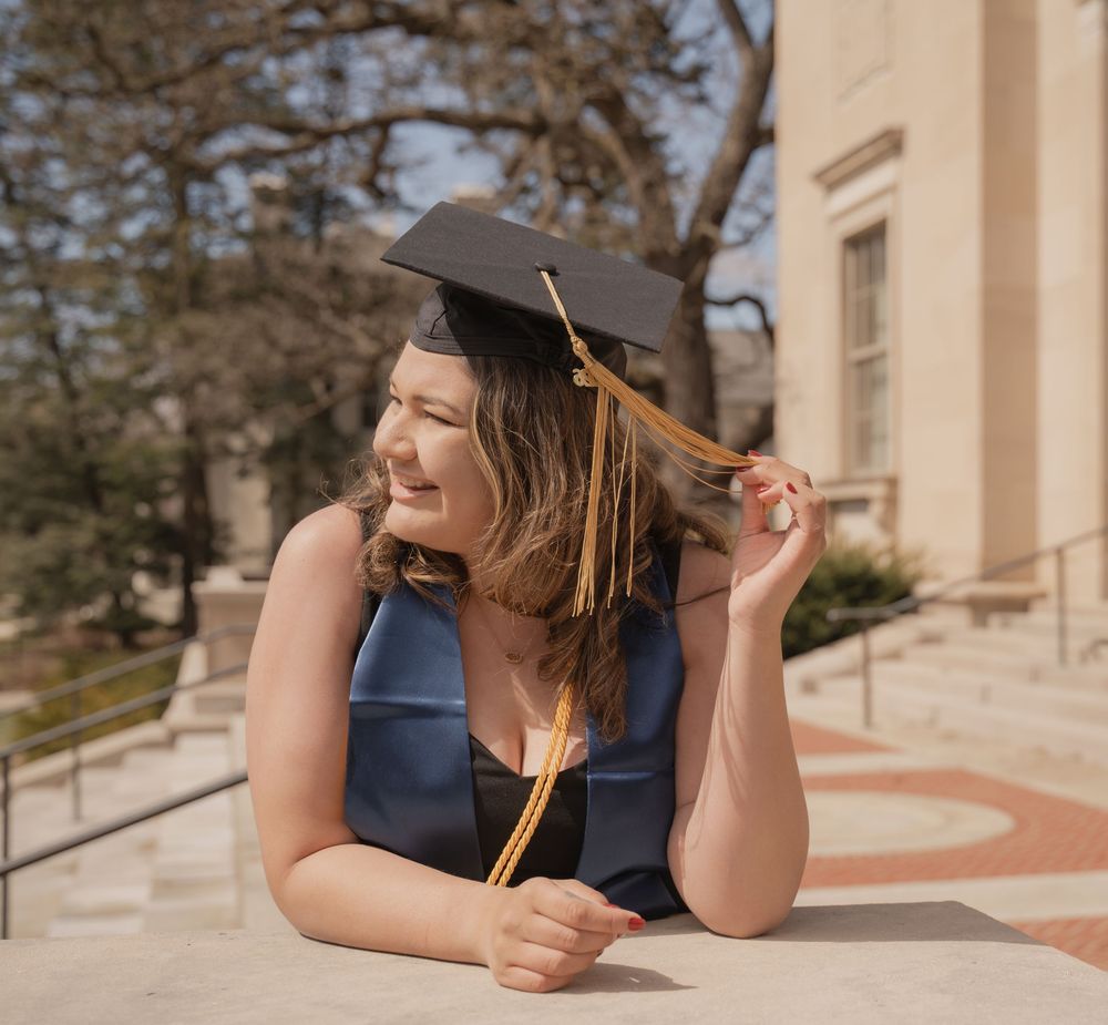 Smiling female graduate in cap and gown poses outdoors on college campus steps on a sunny day.