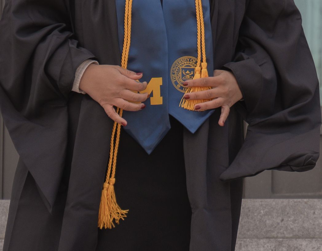 Graduate in black cap and gown wearing gold honor cords and blue University of Michigan stole at graduation ceremony.