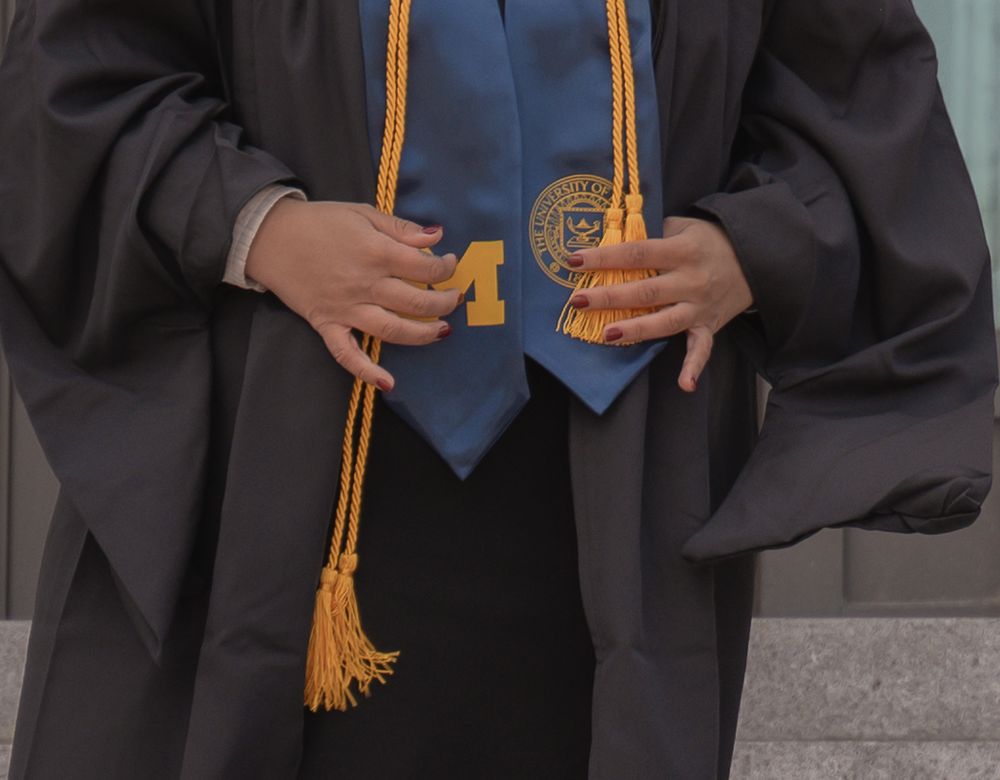 Graduate in black cap and gown wearing gold honor cords and blue University of Michigan stole at graduation ceremony.