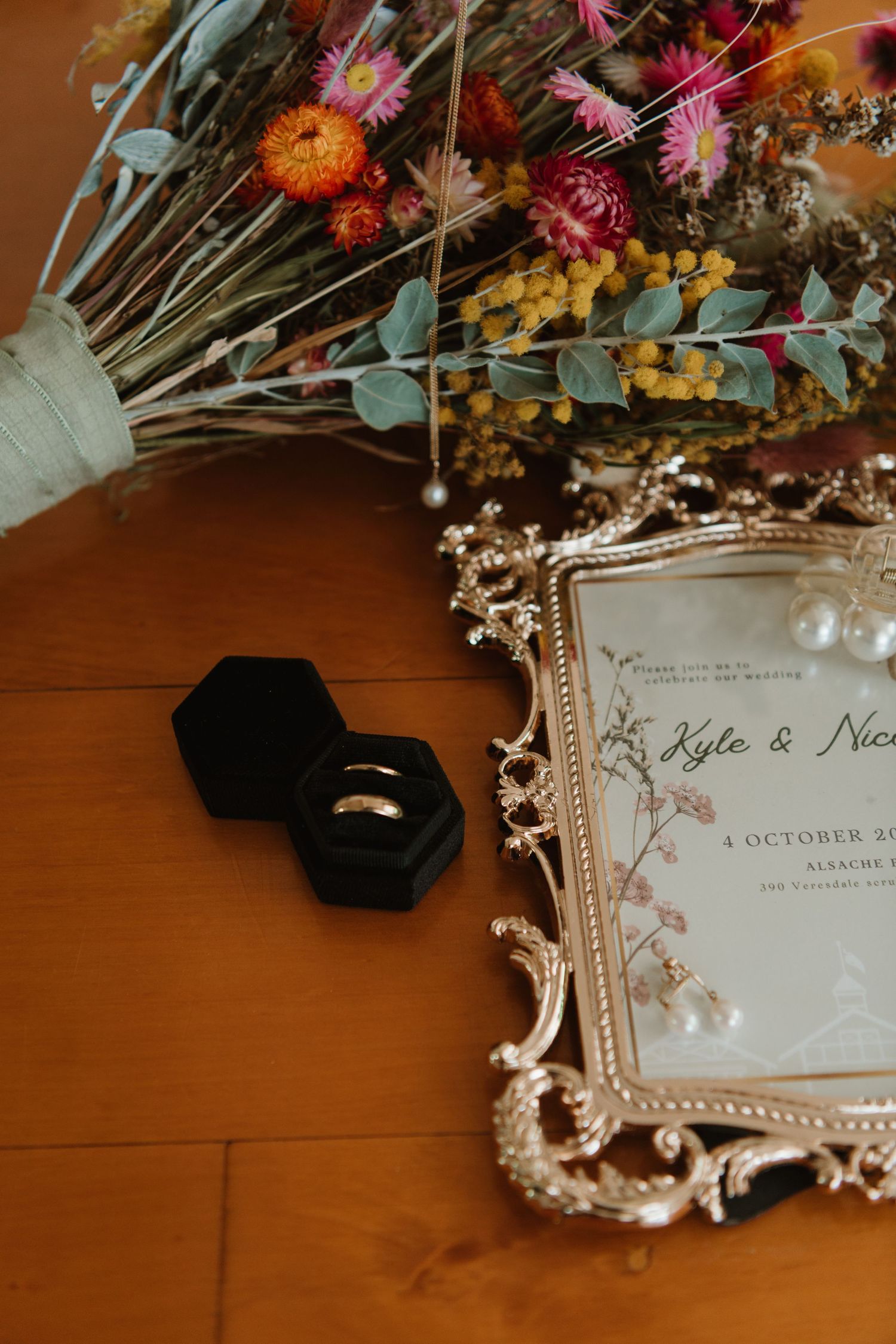 Wedding rings in velvet box beside ornate gold-framed invitation and dried floral bouquet on wooden surface.