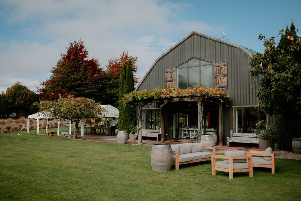 Outdoor wedding venue with rustic barn buildings, wooden benches, and lush green landscaping in autumn setting at Lookout Lodge Wanaka, New