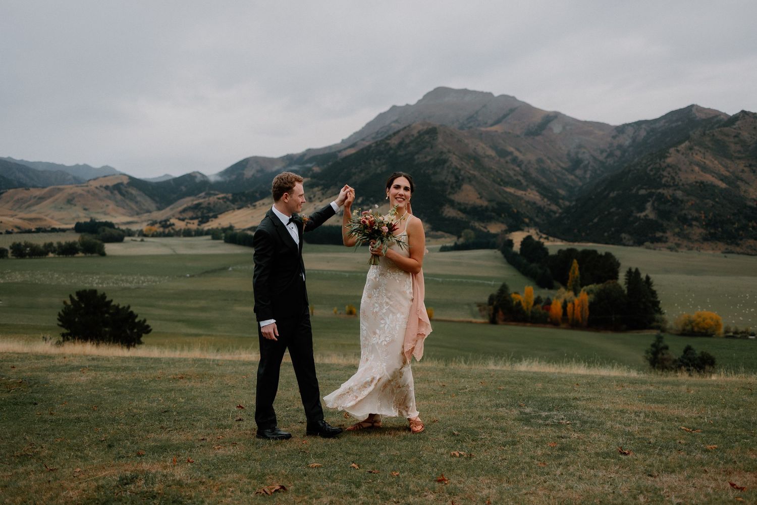 A couple celebrates their wedding day against a backdrop of rolling mountains and golden fields at sunset at Lookout Lodge Wanaka.