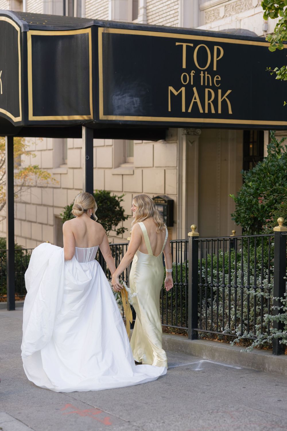 Wedding party members in white dresses and formal attire walk along a covered walkway with elegant black awnings.