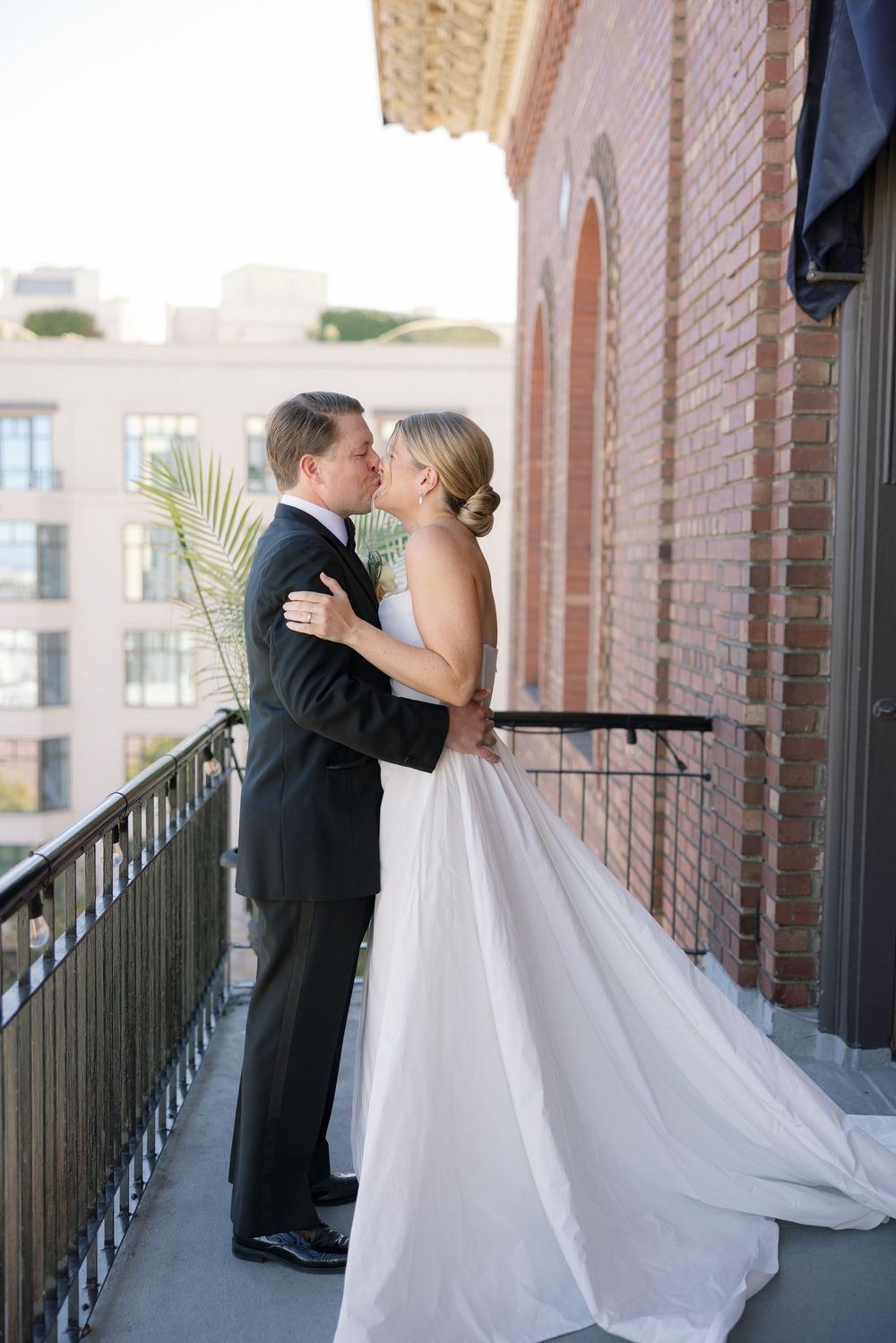 A romantic wedding couple shares a kiss on a brick balcony with a flowing white dress and classic black tuxedo.