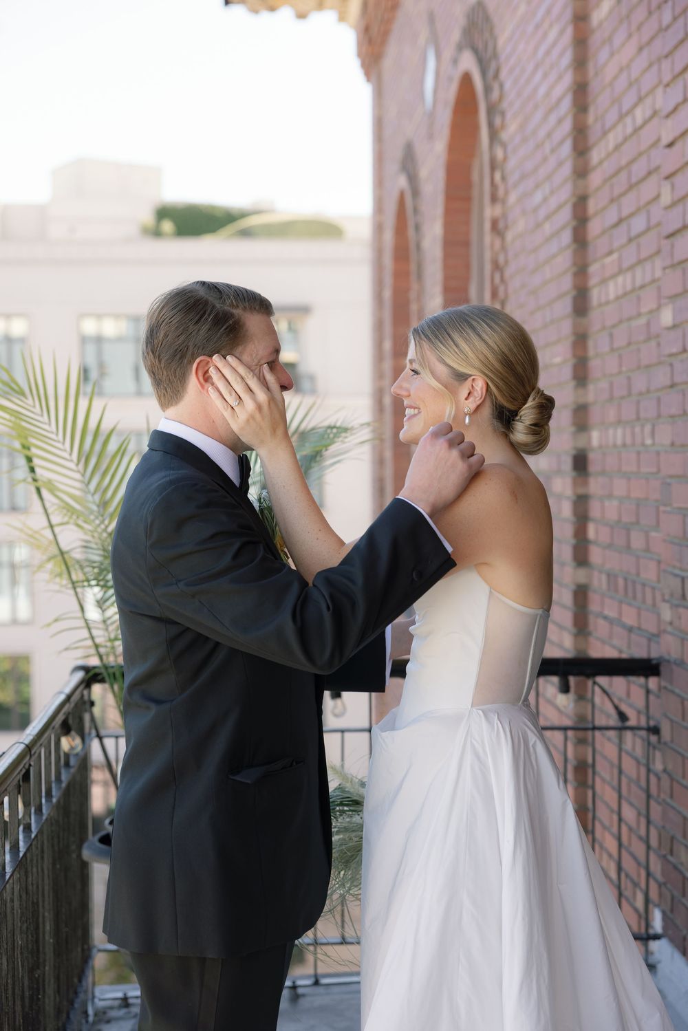 A tender moment between a couple in wedding attire on a brick balcony with palm fronds.