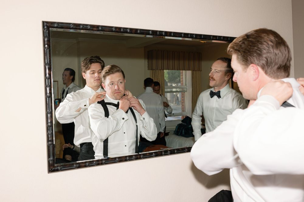 Someone in a white tuxedo jacket adjusts their bowtie while looking in a mirror during formal preparations.
