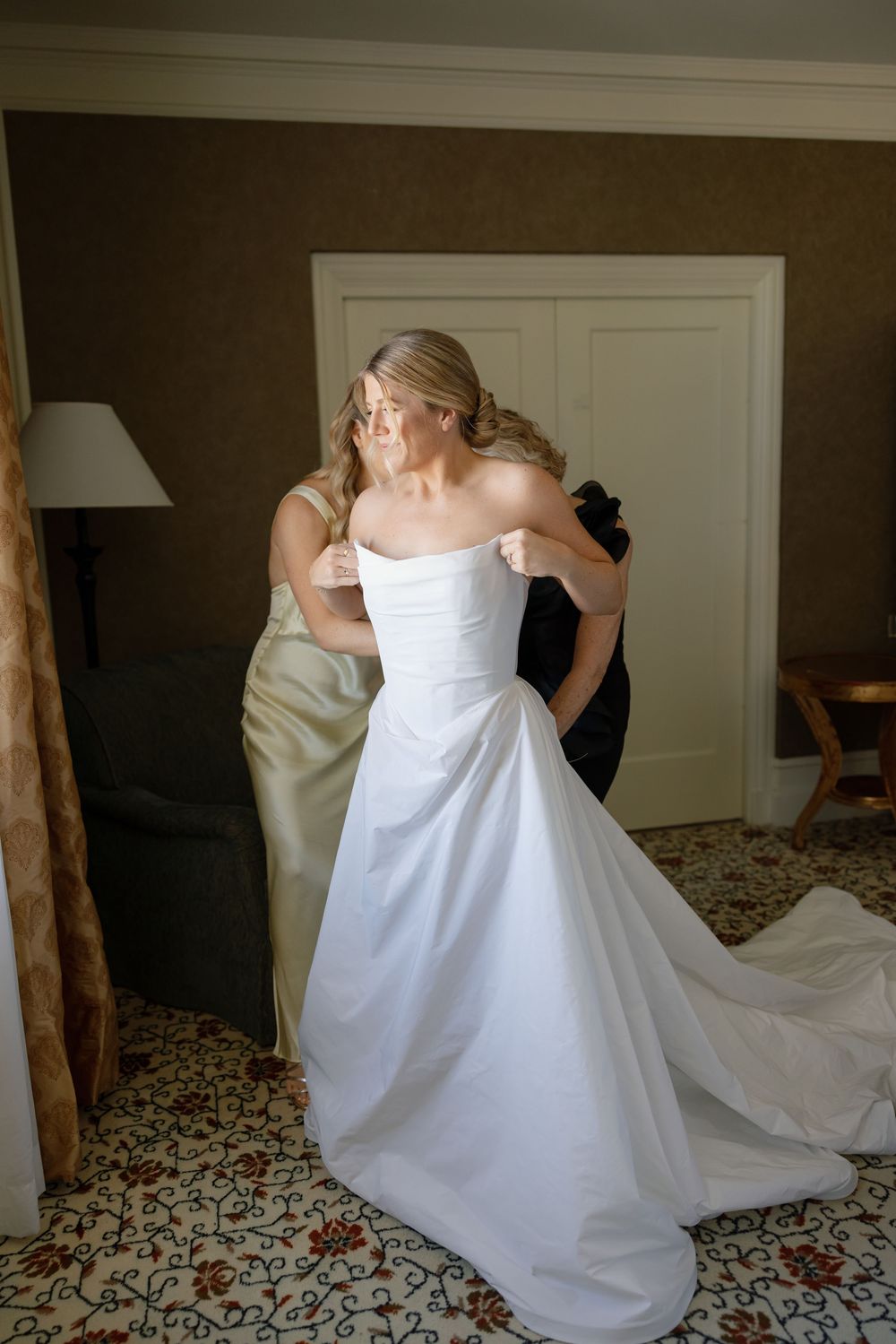 A bride in an elegant white strapless wedding gown adjusts her dress in a room with patterned carpet and warm lighting.