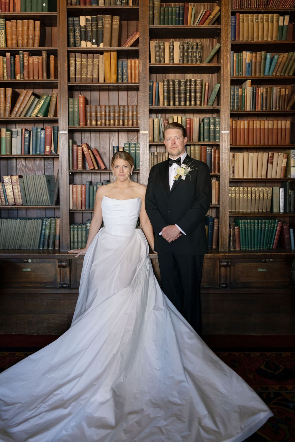 A wedding couple poses in front of a grand library wall filled with vintage books and wooden shelving.
