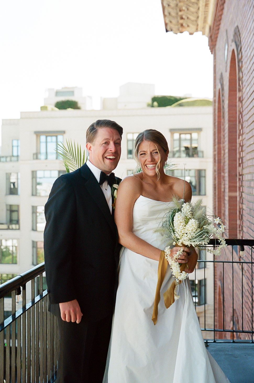 A joyful wedding couple shares candid moments on a balcony in formal attire.