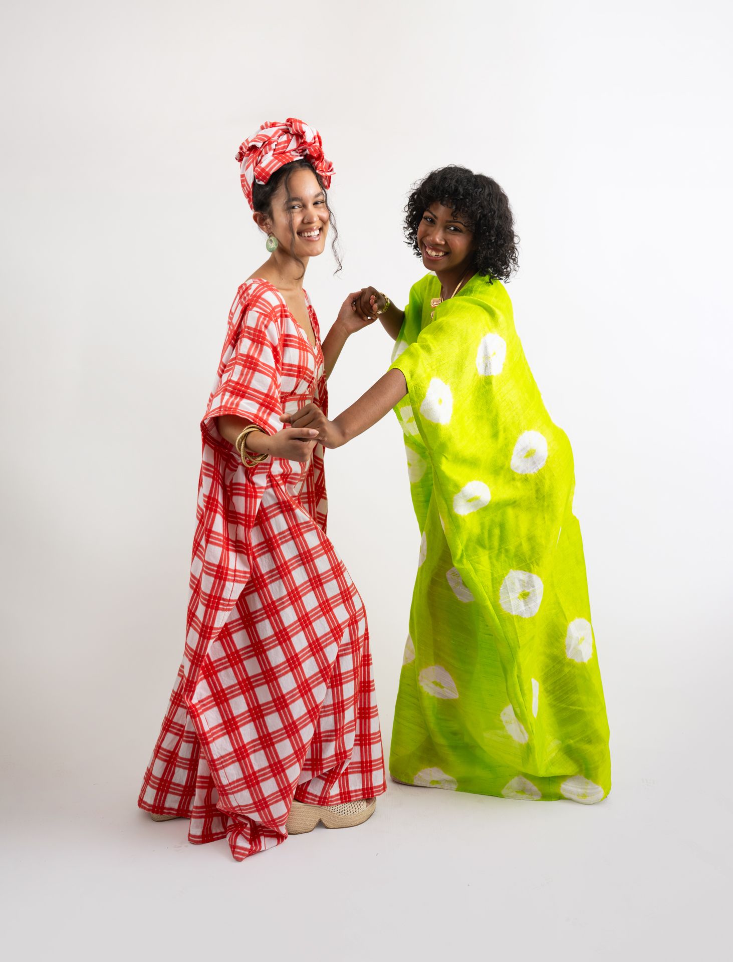 Two women smiling in vibrant African-inspired maxi dresses, one red gingham with head wrap, one lime green with floral print.