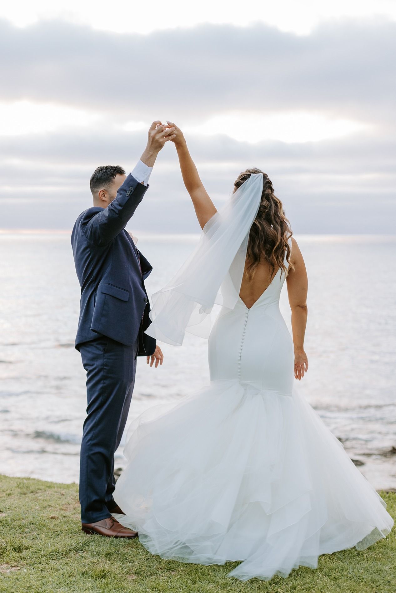 Bride and groom dancing by the ocean, bride in backless mermaid gown with veil, groom in navy suit in La Jolla.