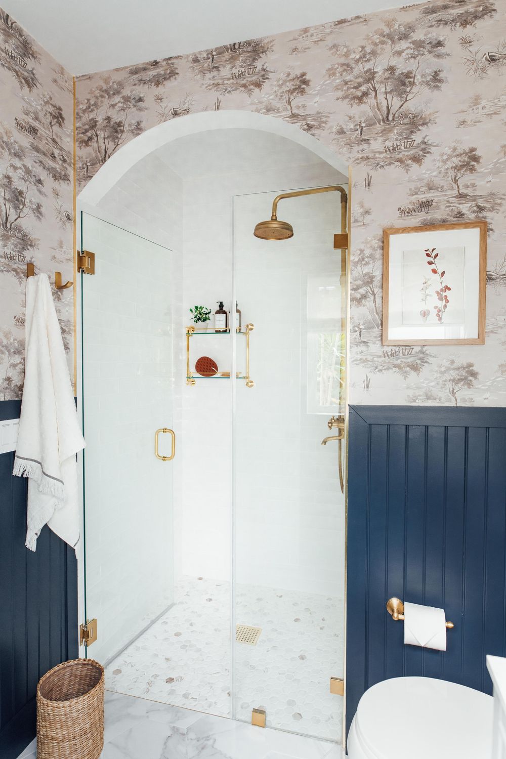 Elegant bathroom with brass shower fixtures, navy wainscoting, toile wallpaper, and glass shower door.
