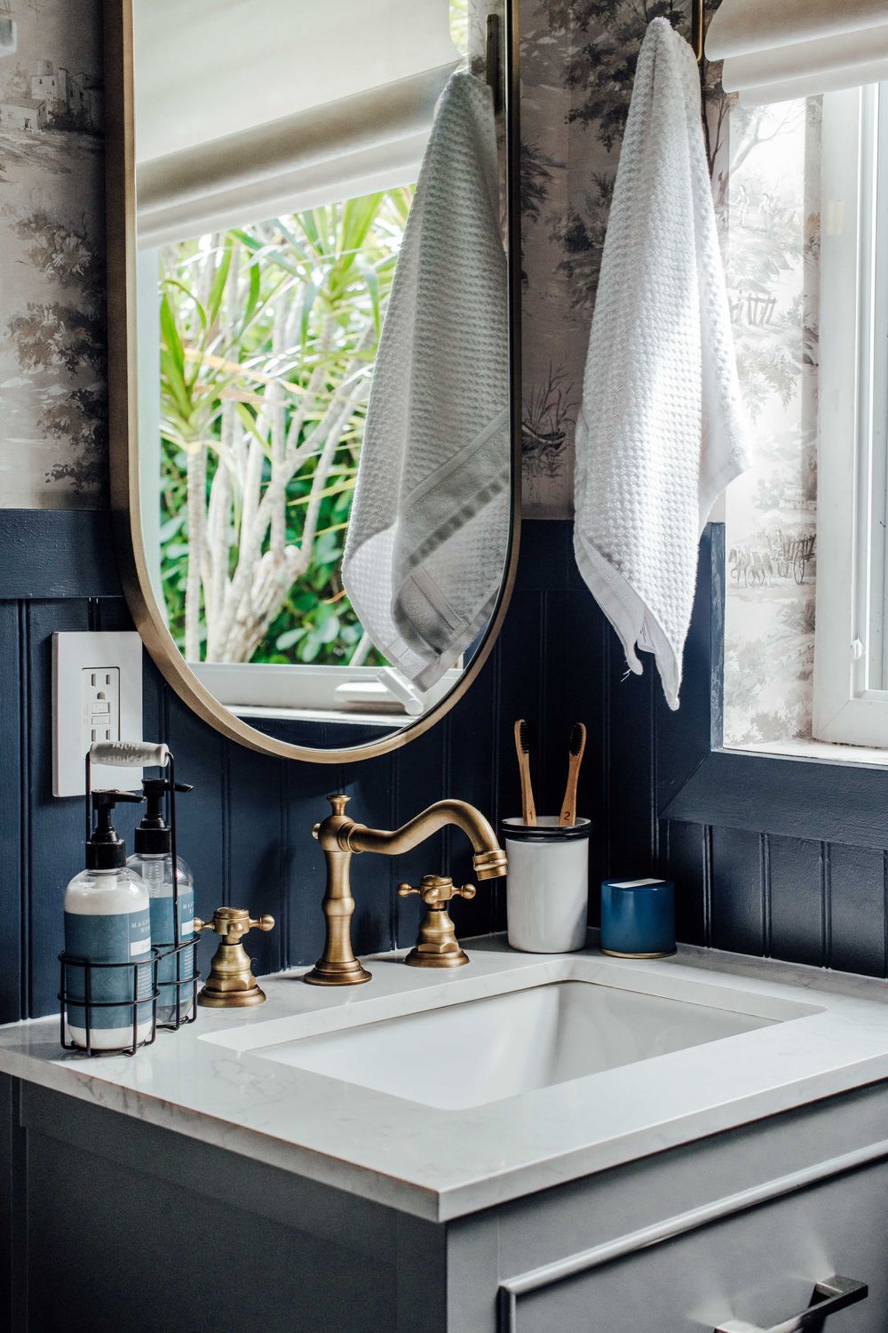 Navy blue bathroom vanity with brass faucet, oval mirror, marble countertop, and toile wallpaper.
