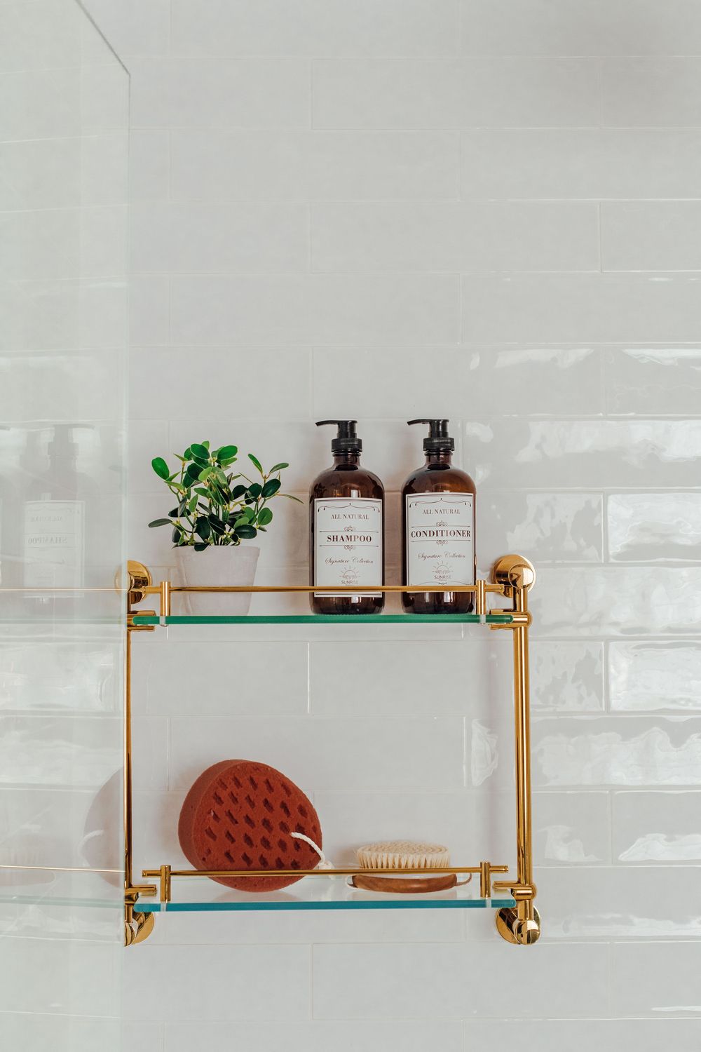 Gold brass shower shelf with luxury shampoo bottles, small plant, and wooden brush on white tiles.