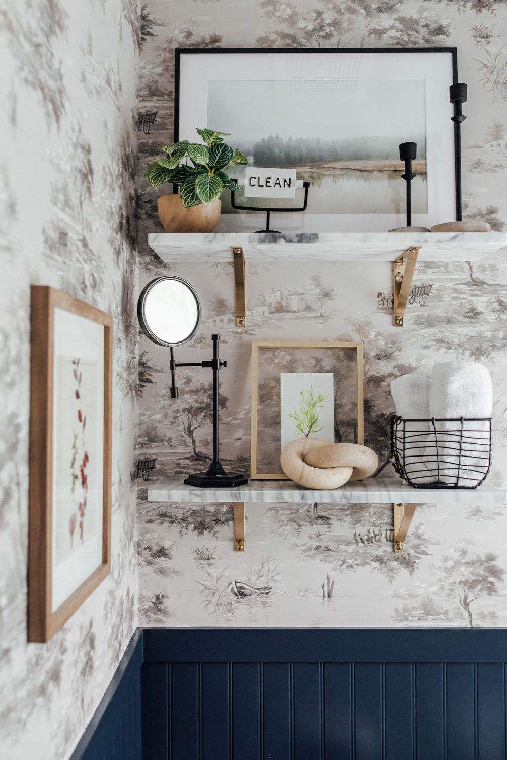 Bathroom shelves with toile wallpaper, navy wainscoting, decorative mirror, plants, and framed art.