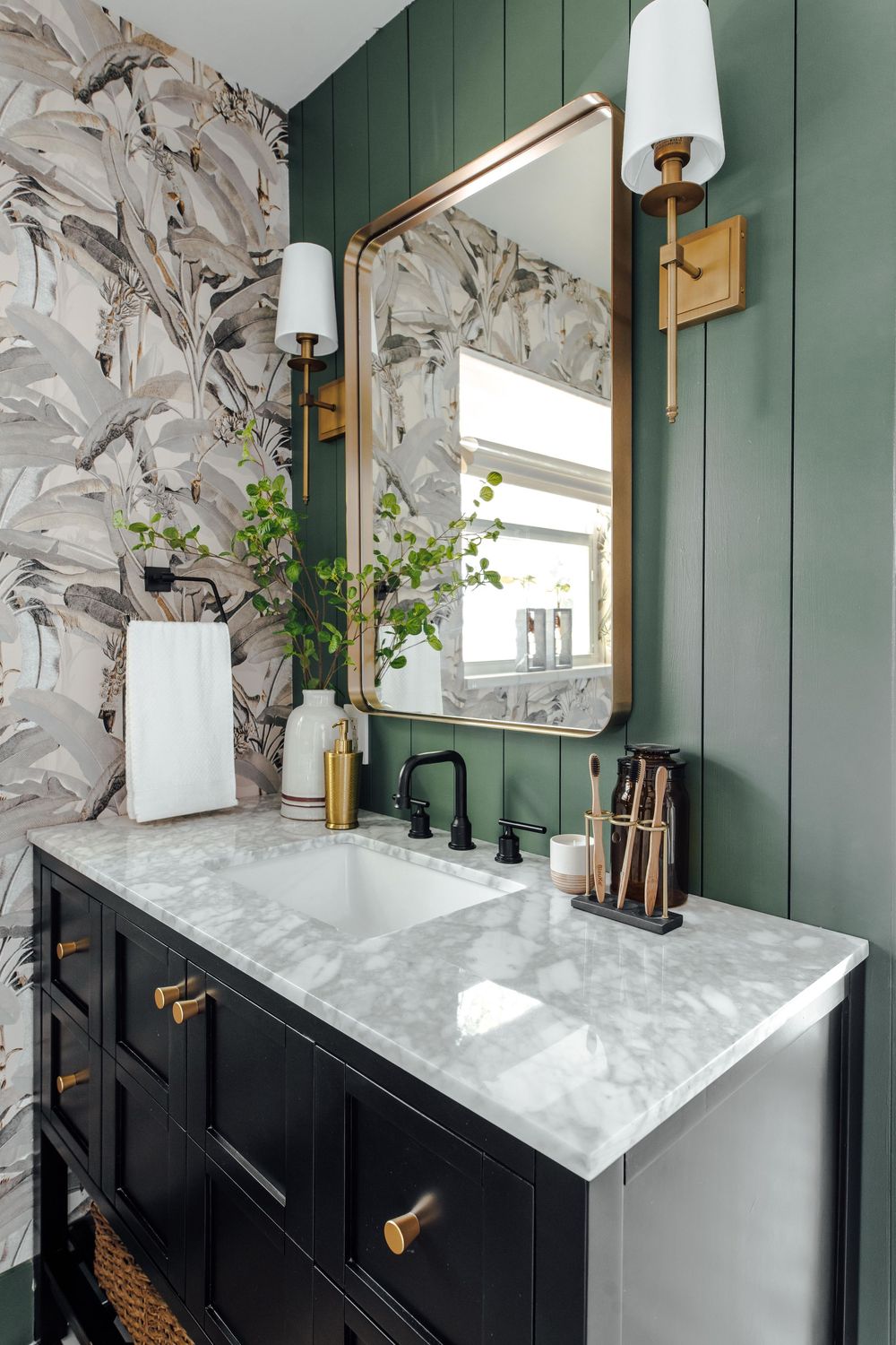 Black bathroom vanity with marble countertop, brass mirror, green shiplap wall, and bold wallpaper.
