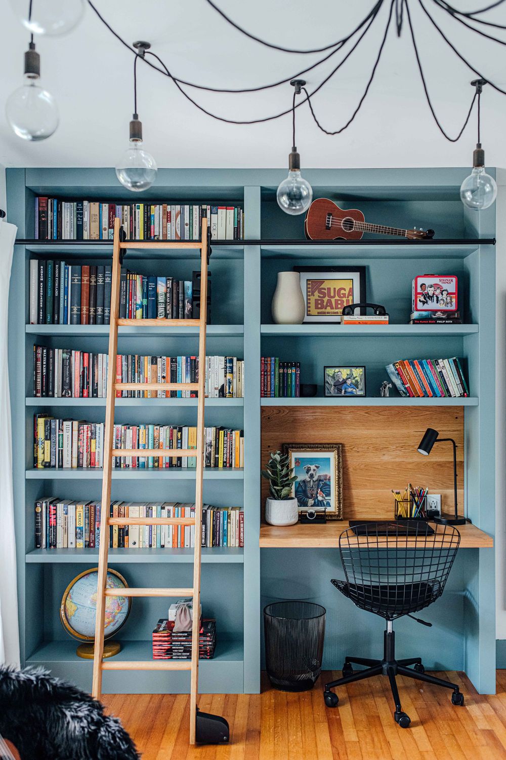 Blue built-in bookcase with wooden ladder, desk, books, guitar, and Edison bulb pendant lights.