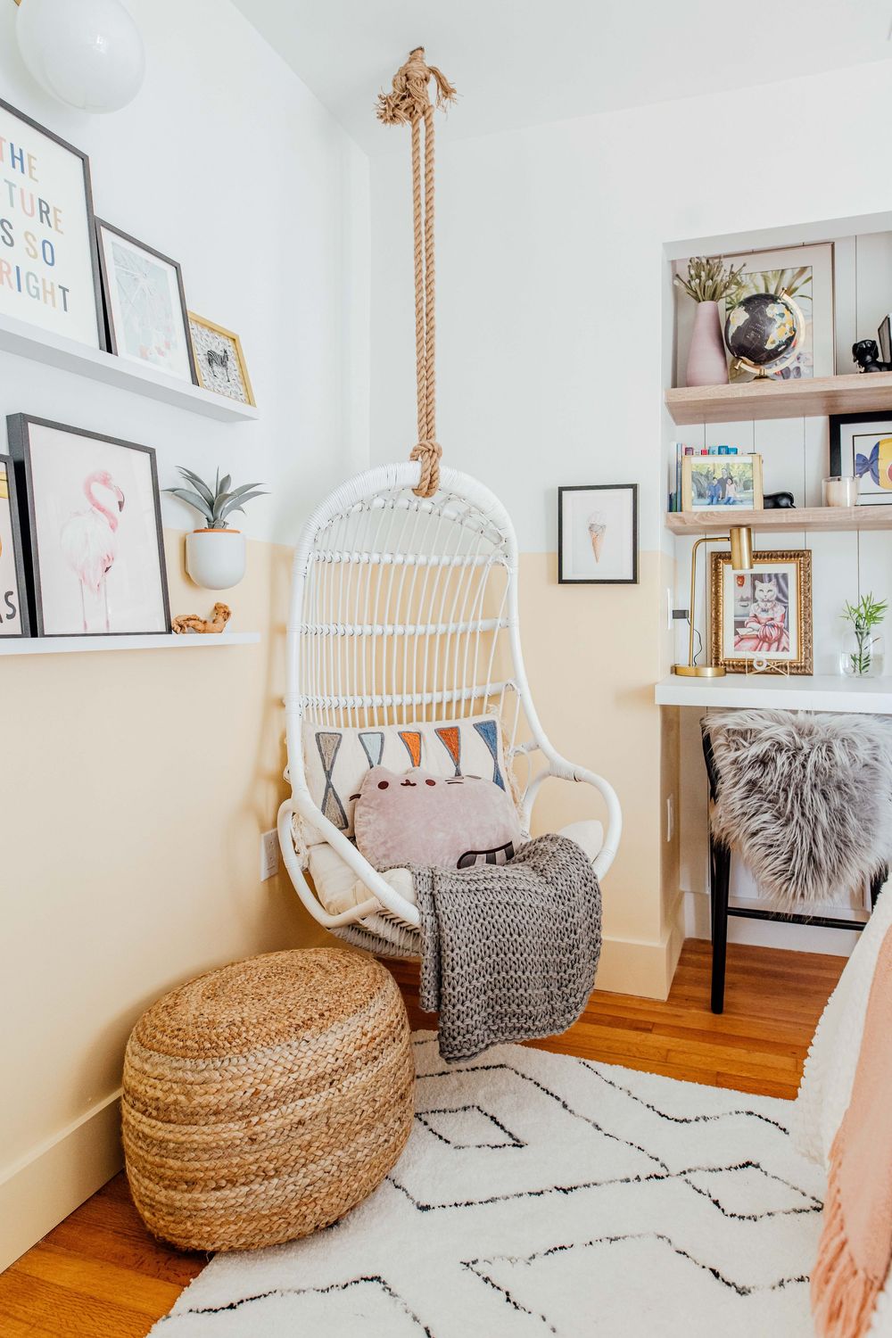 White rattan hanging chair with woven pouf on rug beside gallery wall in cozy bedroom.