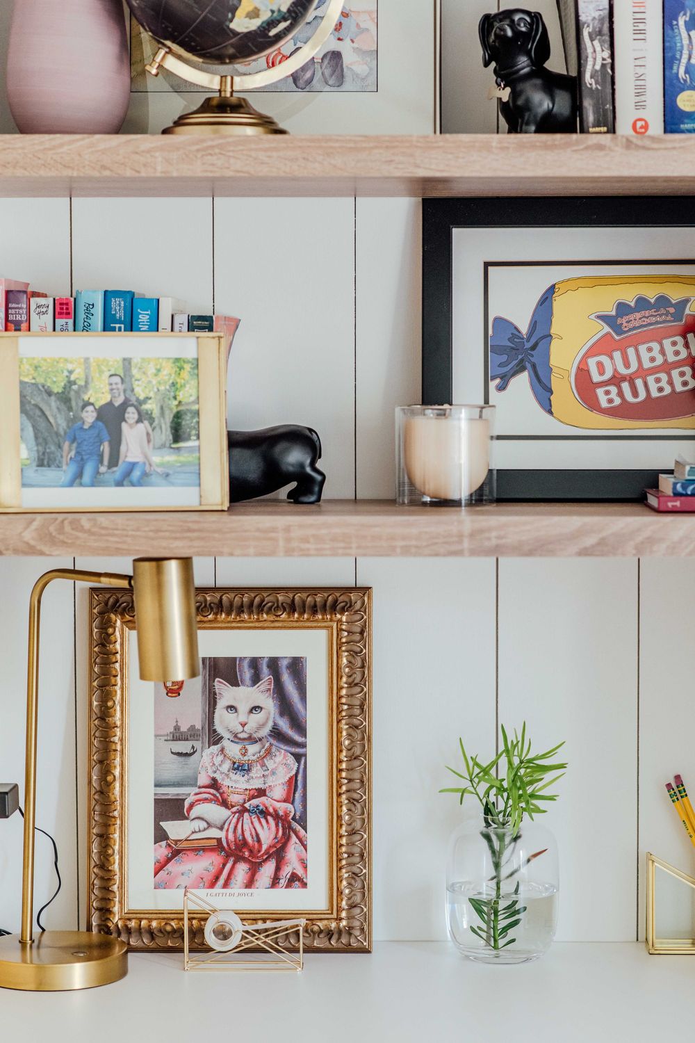 Desk shelf styled with gold lamp, framed cat art, family photo, globe, and Dubble Bubble art.