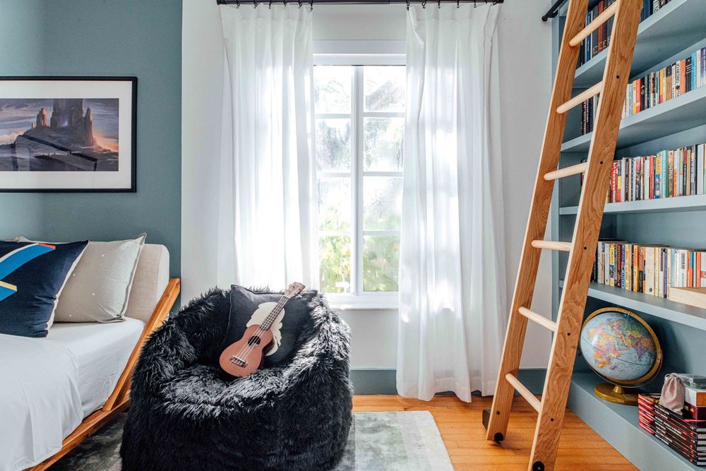 Blue bedroom with bean bag chair, ukulele, wooden ladder bookshelf, white curtains, and framed landscape wall art.