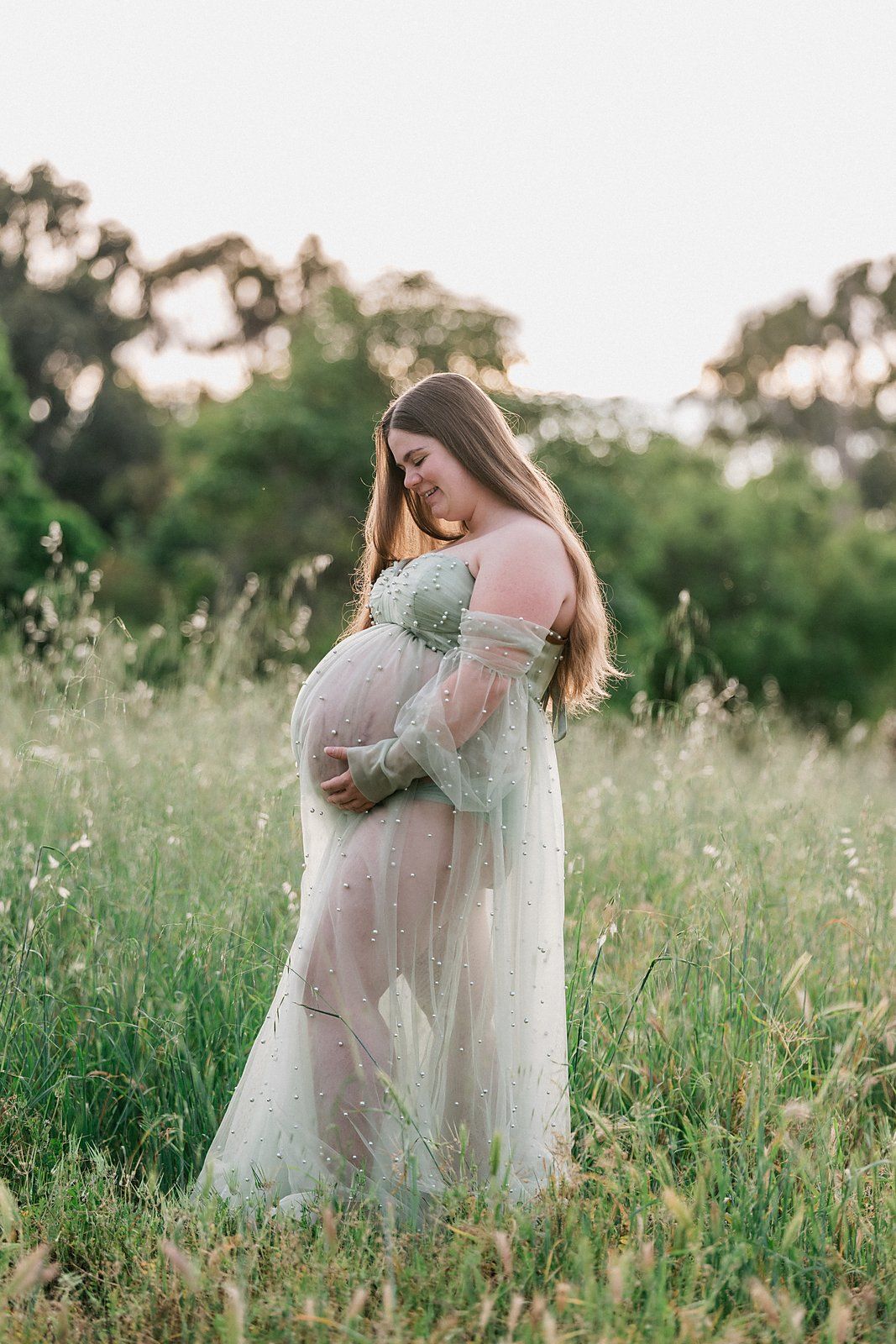 Pregnant woman in sheer floral gown cradling her baby bump in a lush green meadow at golden hour.