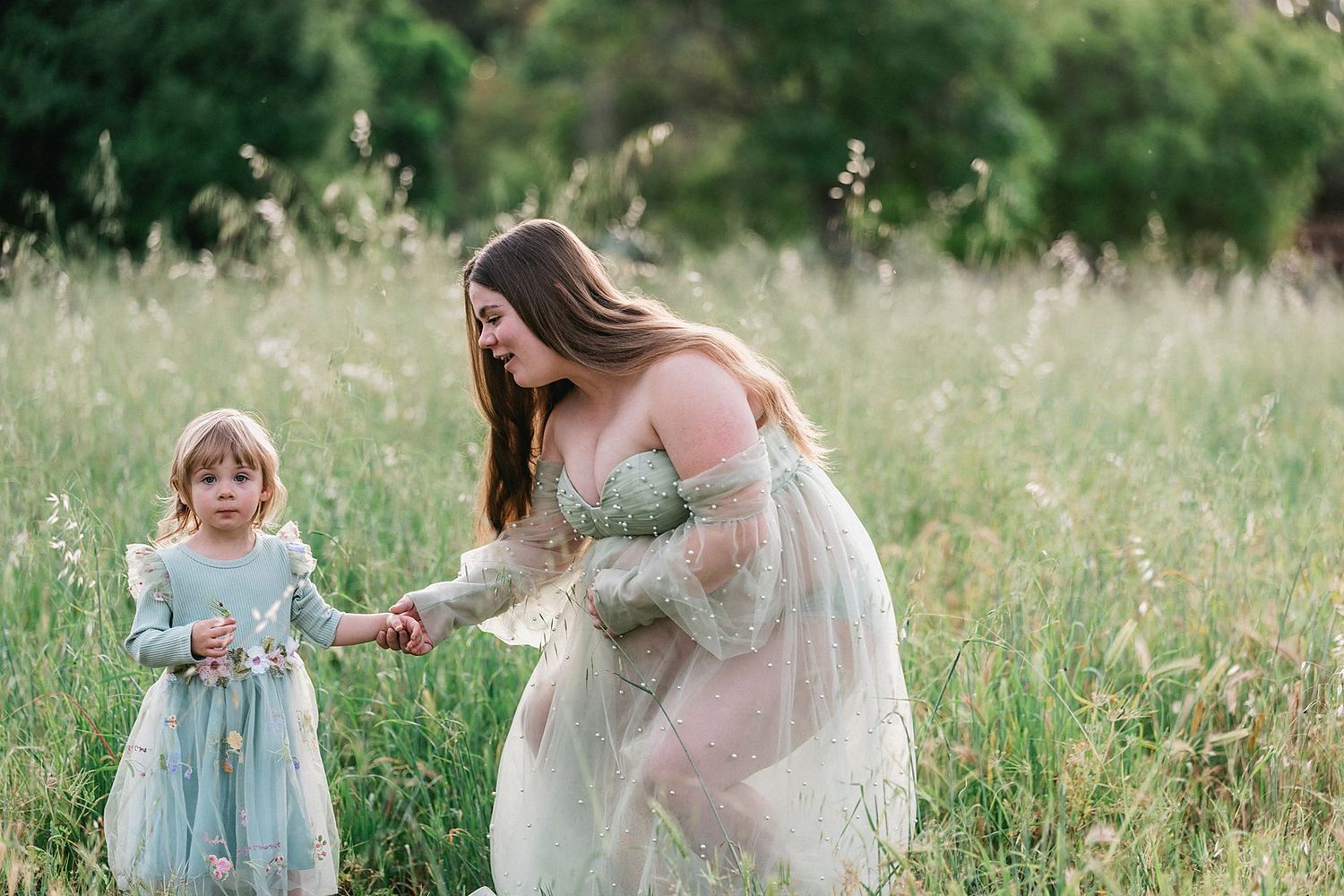 A pregnant woman in a sheer gown holds hands with a young girl in a blue dress in a lush green meadow.