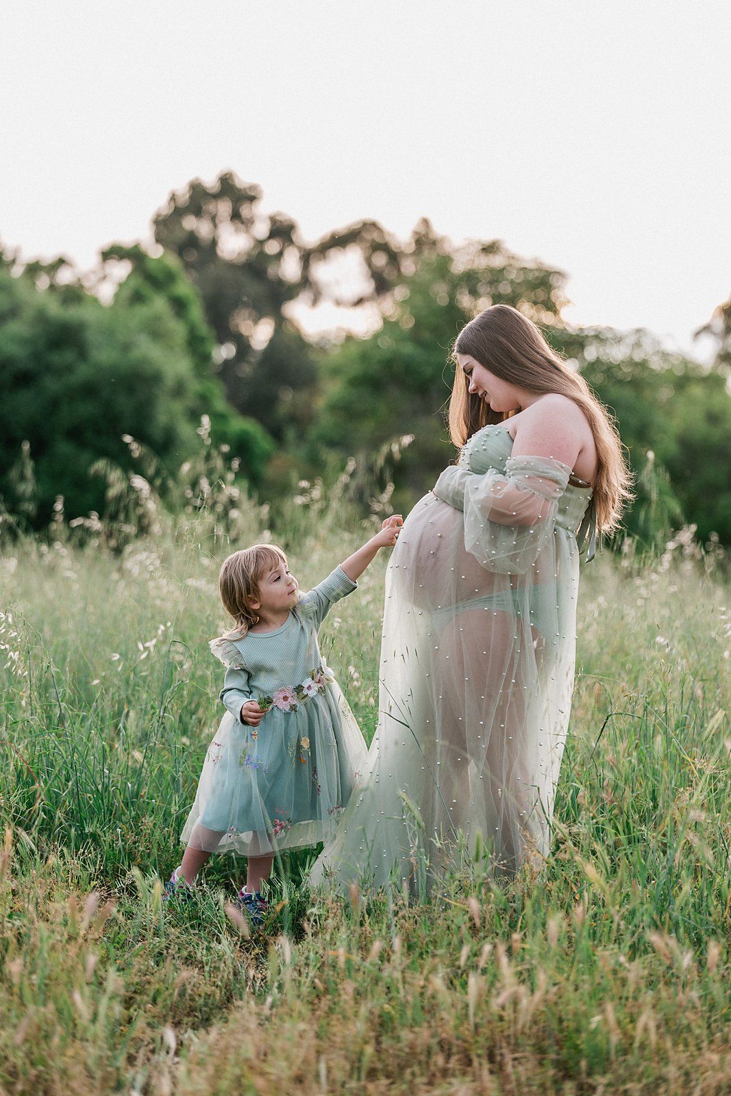 Pregnant woman in sheer green gown with young daughter in field during golden hour maternity photoshoot.