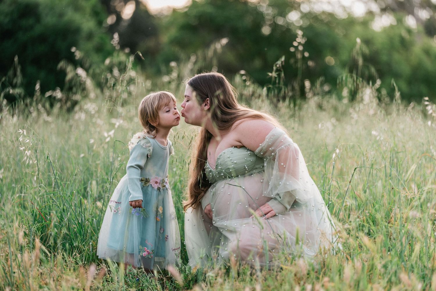 Pregnant woman in green tulle gown kissing toddler girl in floral dress in a sunlit meadow.