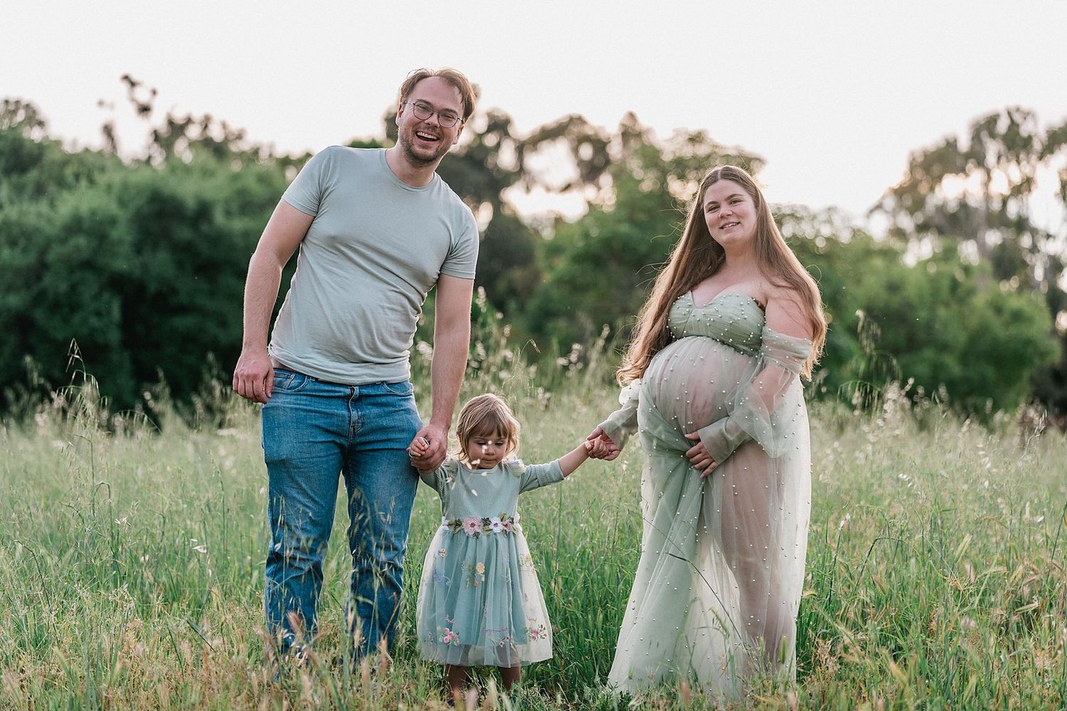 Pregnant woman in green dress with husband and toddler daughter posing in a sunny meadow for maternity photo.