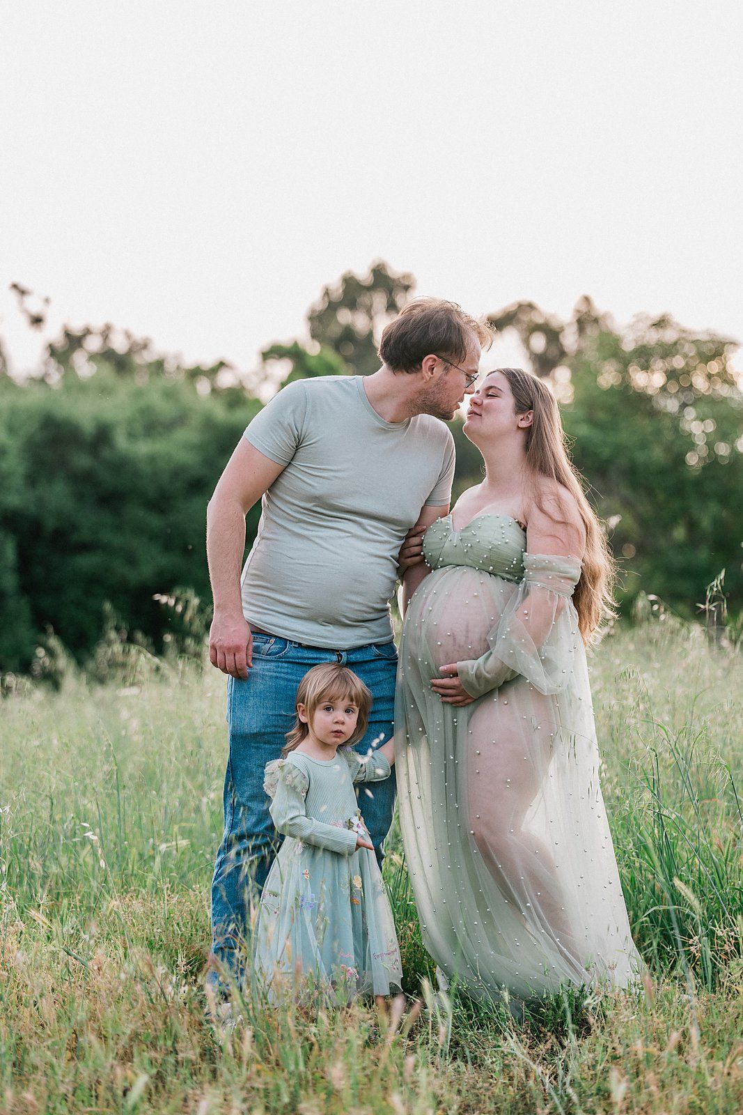 Pregnant woman in sheer gown kissing man in field while toddler girl stands nearby during maternity photoshoot.