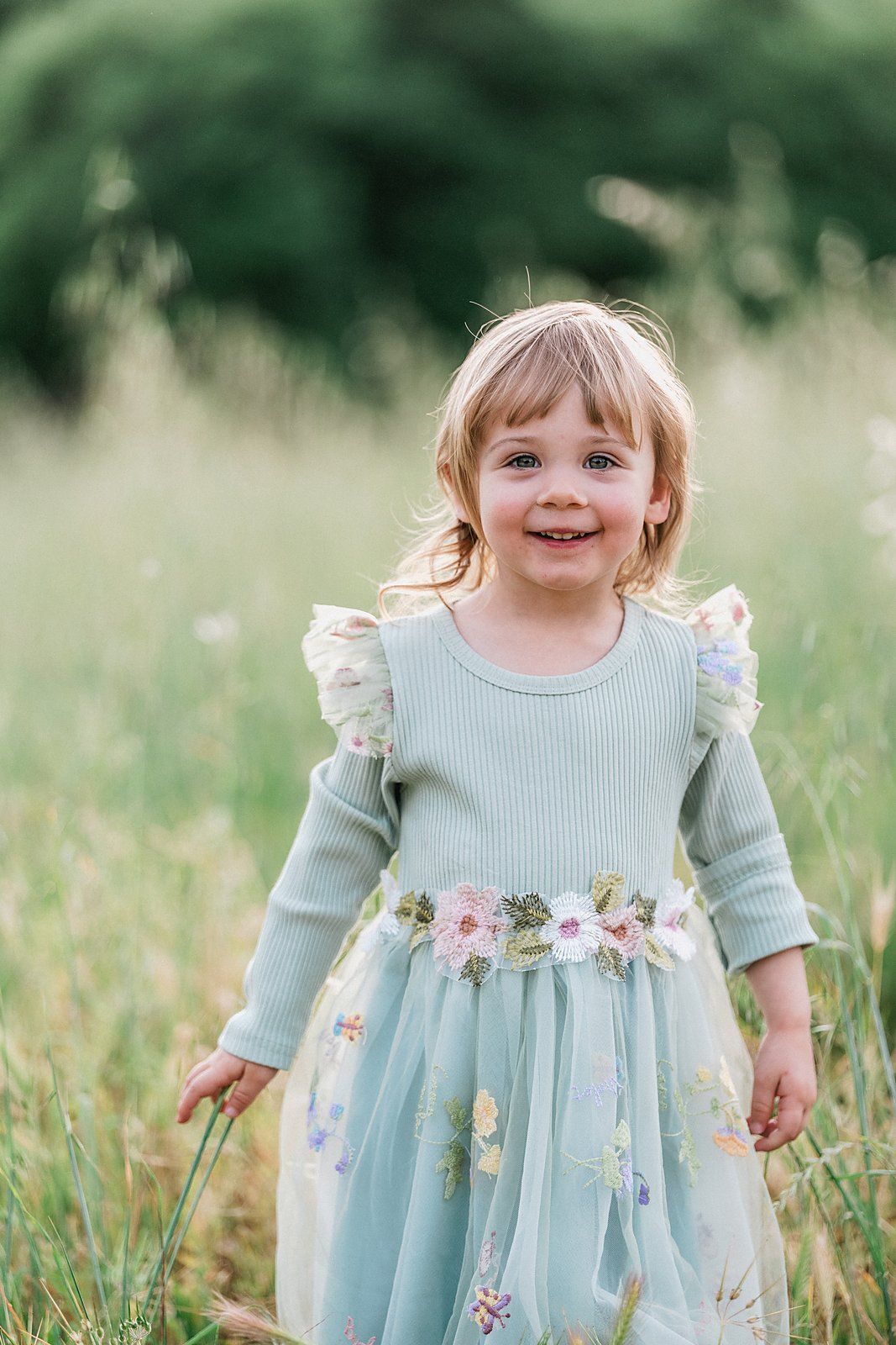 A smiling toddler girl in a mint green floral embroidered dress stands in a lush green meadow field.