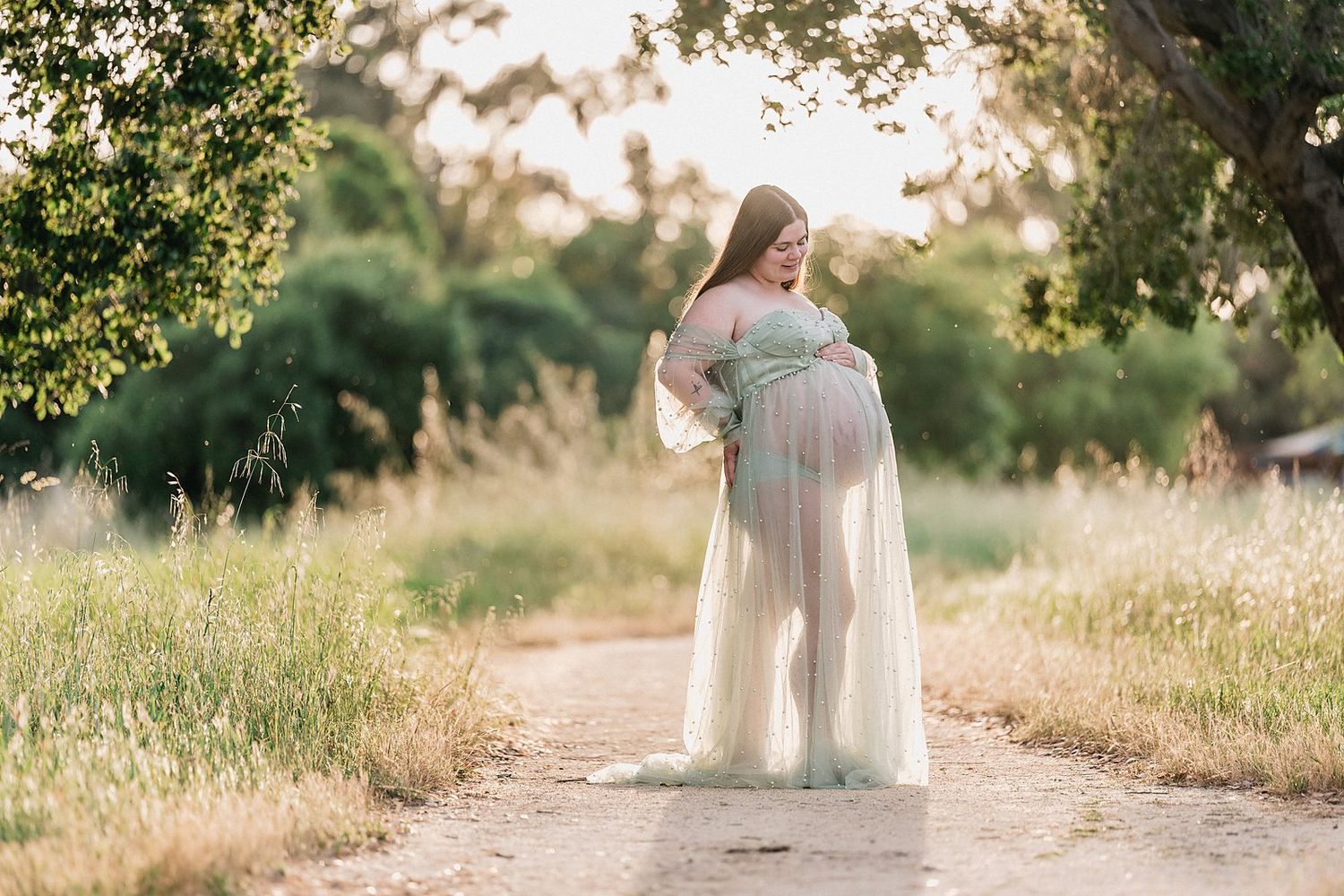 Pregnant woman in sheer white maternity gown standing on a sunlit dirt path surrounded by lush green trees.